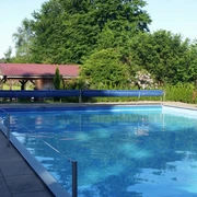 Freibad Das Bild zeigt ein Freibad mit einem großen rechteckigen Pool, umgeben von Bäumen und einer Wiese. Im Hintergrund ist ein kleines Gebäude mit rotem Dach zu sehen. Links vom Bild sieht man einen Kinderspielplatz mit bunten Spielgeräten. Diese erholsame Umgebung lädt zum Entspannen und Spielen ein.