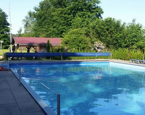 Freibad Das Bild zeigt ein Freibad mit einem großen rechteckigen Pool, umgeben von Bäumen und einer Wiese. Im Hintergrund ist ein kleines Gebäude mit rotem Dach zu sehen. Links vom Bild sieht man einen Kinderspielplatz mit bunten Spielgeräten. Diese erholsame Umgebung lädt zum Entspannen und Spielen ein.