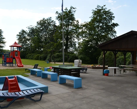 Spielplatz  Ein Spielplatz mit Rutsche, Wipp-Tier, Picknickbänken und überdachtem Essbereich in einer grünen Umgebung.