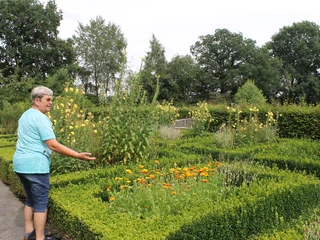 KneippGarten Eine Person in türkisfarbener Kleidung steht im Grünen, umgeben von bunten Blumenbeeten und hohen Büschen.