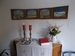 Kapelle "Unter den Linden" Ein Altar in der Kapelle mit zwei brennenden Kerzen, Blumenstrauß, Bibel und bemalten Bildern.