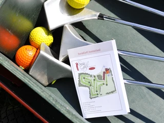 Ausstattung Swingolf Vier Golfschläger und farbige Golfbälle in einem Transportwagen, daneben eine Karte der Golfanlage.