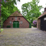 Heinrich Kunst Haus Das Bild zeigt einen kleinen idyllischen Hof umgeben von Bäumen. Im Zentrum steht ein traditionelles rotes Backsteinhaus mit grünem Holztor und kleinen runden Fenstern. Rechts daneben befindet sich ein dunkles Fachwerkgebäude. Die gepflasterte Fläche und das grüne Blattwerk erzeugen eine ruhige, einladende Atmosphäre. Dieses Setting vermittelt den Charme ländlichen Lebens und Ruhe.