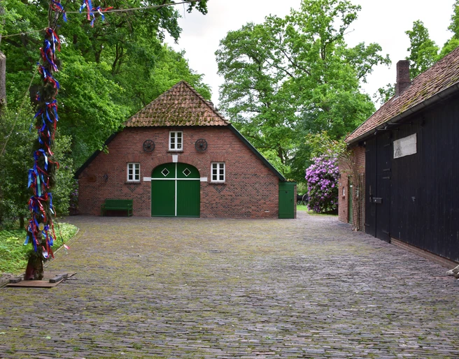 Heinrich Kunst Haus Das Bild zeigt einen kleinen idyllischen Hof umgeben von Bäumen. Im Zentrum steht ein traditionelles rotes Backsteinhaus mit grünem Holztor und kleinen runden Fenstern. Rechts daneben befindet sich ein dunkles Fachwerkgebäude. Die gepflasterte Fläche und das grüne Blattwerk erzeugen eine ruhige, einladende Atmosphäre. Dieses Setting vermittelt den Charme ländlichen Lebens und Ruhe.