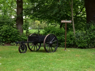 Heinrich Kunst Haus Ein alter Karren steht auf einer Wiese. Dahinter sind dichte grüne Bäume. Ein Schild ist daneben.