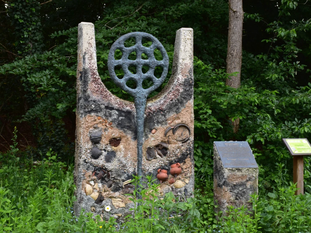 Hügelgrab Ein modernes Kunstwerk aus Stein, markiert und verziert mit Kreisen, Flaschen und kleinen Töpfen, steht vor einem bewaldeten Hintergrund.