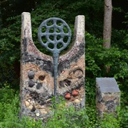 Hügelgrab Ein modernes Kunstwerk aus Stein, markiert und verziert mit Kreisen, Flaschen und kleinen Töpfen, steht vor einem bewaldeten Hintergrund.