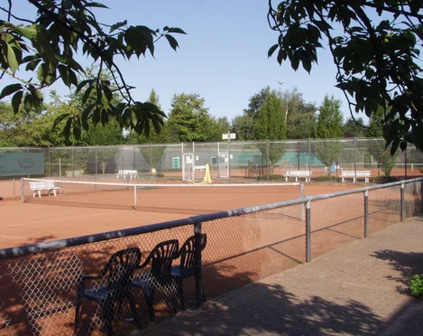 Tennisplatz Tennisplatz mit rotem Sand, umgeben von Bäumen, Zaun im Vordergrund, leere Bänke am Rand.