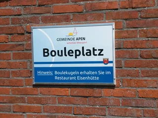 Platzschild Das Bild zeigt ein Schild mit der Aufschrift "Gemeinde Apen - Natürlich lebenswert - Bouleplatz". Unten steht der Hinweis, dass Boulekugeln im Restaurant Eisenhütte erhältlich sind. Das Schild ist an einer roten Backsteinwand angebracht.