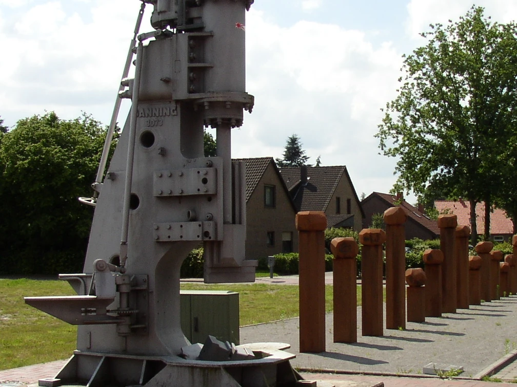Industriedenkmal_Eisenhüttenplatz Industriedenkmal mit historischem Hammerwerk, eingerahmt von Bäumen und Häusern im Hintergrund.