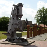 Industriedenkmal_Eisenhüttenplatz Industriedenkmal mit historischem Hammerwerk, eingerahmt von Bäumen und Häusern im Hintergrund.