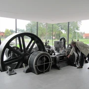 Schauraum Stahlwerk Augustfehn Im Schauraum des Stahlwerks Augustfehn zeigt das Bild eine historische Dampfmaschine mit großen, schwarzen Schwungrädern in einem modernen Ausstellungsraum mit Glaswand. Menschen stehen interessiert um die Maschine.