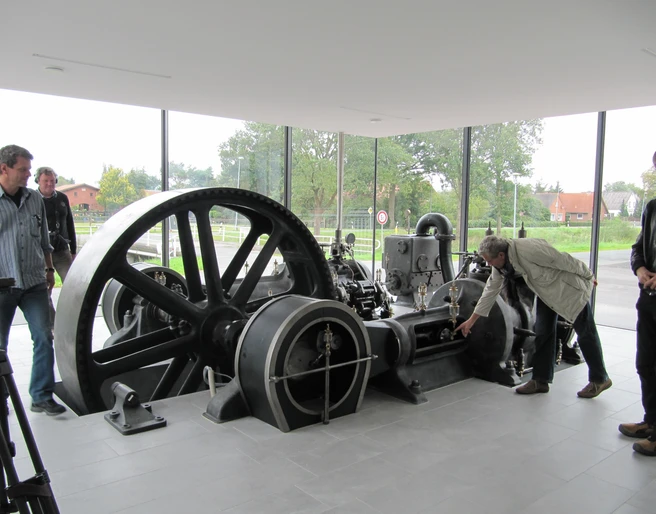 Schauraum Stahlwerk Augustfehn Im Schauraum des Stahlwerks Augustfehn zeigt das Bild eine historische Dampfmaschine mit großen, schwarzen Schwungrädern in einem modernen Ausstellungsraum mit Glaswand. Menschen stehen interessiert um die Maschine.