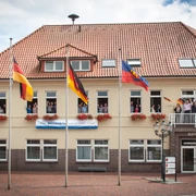 Rathaus_Gemeinde Apen Das Rathaus der Gemeinde Apen, ein zweigeschossiges Gebäude mit rotem Ziegeldach, ist geschmückt mit Blumenkästen und mehreren Flaggenmasten. Vor dem Gebäude stehen Fahnen, inklusive der deutschen und der europäischen Flagge, im leichten Wind flatternd.