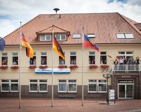 Rathaus_Gemeinde Apen Das Rathaus der Gemeinde Apen, ein zweigeschossiges Gebäude mit rotem Ziegeldach, ist geschmückt mit Blumenkästen und mehreren Flaggenmasten. Vor dem Gebäude stehen Fahnen, inklusive der deutschen und der europäischen Flagge, im leichten Wind flatternd.