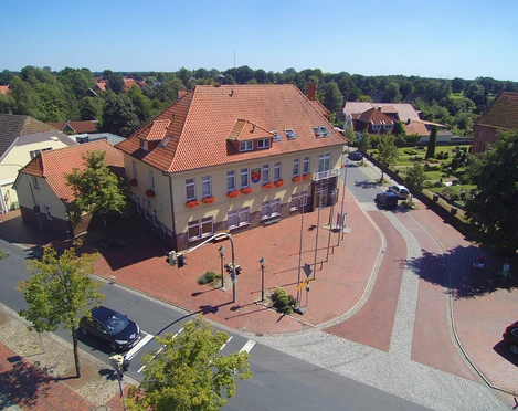 Luftaufnahme_Rathaus_Gemeinde Apen Die Luftaufnahme zeigt das beige Rathaus der Gemeinde Apen mit roten Dachziegeln, umgeben von Bäumen.