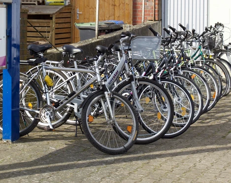 Auswahl der Fahrräder Eine lange Reihe von weißen Fahrrädern steht schräg nebeneinander auf einem gepflasterten Hof. Links ist ein Schild mit der Aufschrift "Fahrradverleih & Fahrradreparatur" sichtbar.