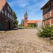 Hippolit Kirche Amelinghausen  Das Bild zeigt einen gepflasterten Weg, flankiert von historischen Fachwerkhäusern, der zur beeindruckenden Hippolit Kirche in Amelinghausen führt. Der Kirchturm erhebt sich majestätisch in den klaren blauen Himmel.