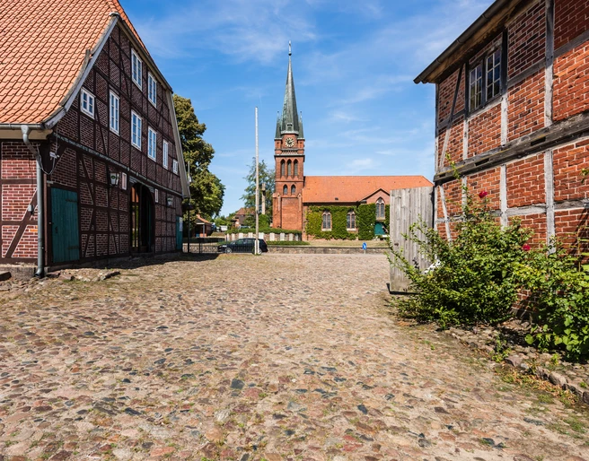 Hippolit Kirche Amelinghausen  Das Bild zeigt einen gepflasterten Weg, flankiert von historischen Fachwerkhäusern, der zur beeindruckenden Hippolit Kirche in Amelinghausen führt. Der Kirchturm erhebt sich majestätisch in den klaren blauen Himmel.