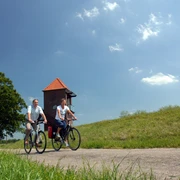 Radlerpaar_Landschaftsfenster_Wasser Ein Radlerpaar fährt bei sonnigem Wetter einen ruhigen Weg entlang, flankiert von grünen Wiesen und einem Baum. Im Hintergrund ist ein gemauerter Turm mit einem roten Dach zu sehen, während leichte Wolken den blauen Himmel zieren.