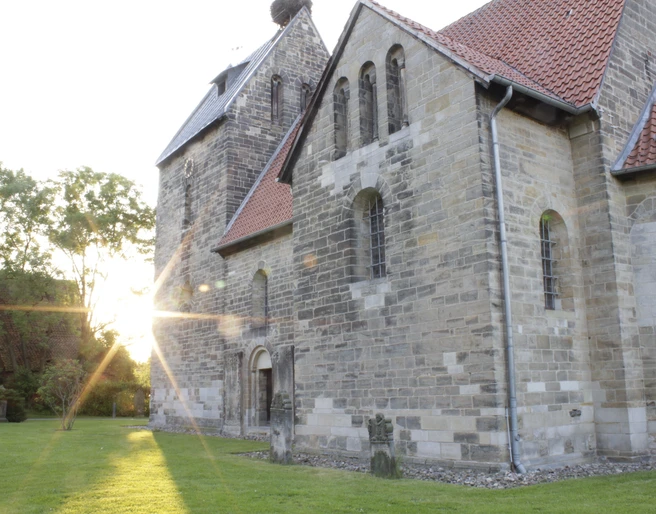 Sigwardskirche Die Sigwardskirche, ein historisches Steingebäude, wird im Abendlicht von der golden untergehenden Sonne angestrahlt, was die Struktur und die Details der gotischen Architektur betont. Links lehnt sich ein Schatten entlang des Rasens, der das friedliche Ambiente untermalt.