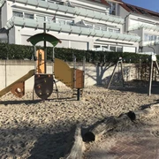 Ein moderner Spielplatz mit Sandboden, einer bunten Rutsche, Schaukel und Klettergerüst vor einer Ferienanlage.