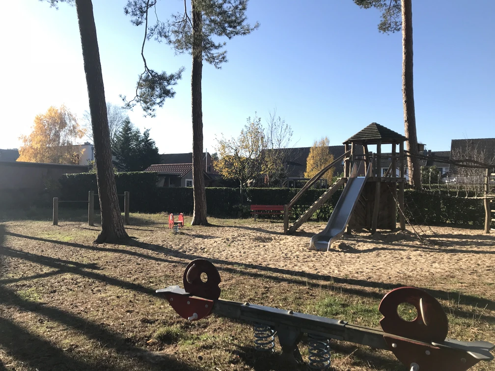 Spielplatz mit Kletterturm, Rutsche und Wipptieren unter hohen Bäumen im herbstlichen Sonnenlicht.