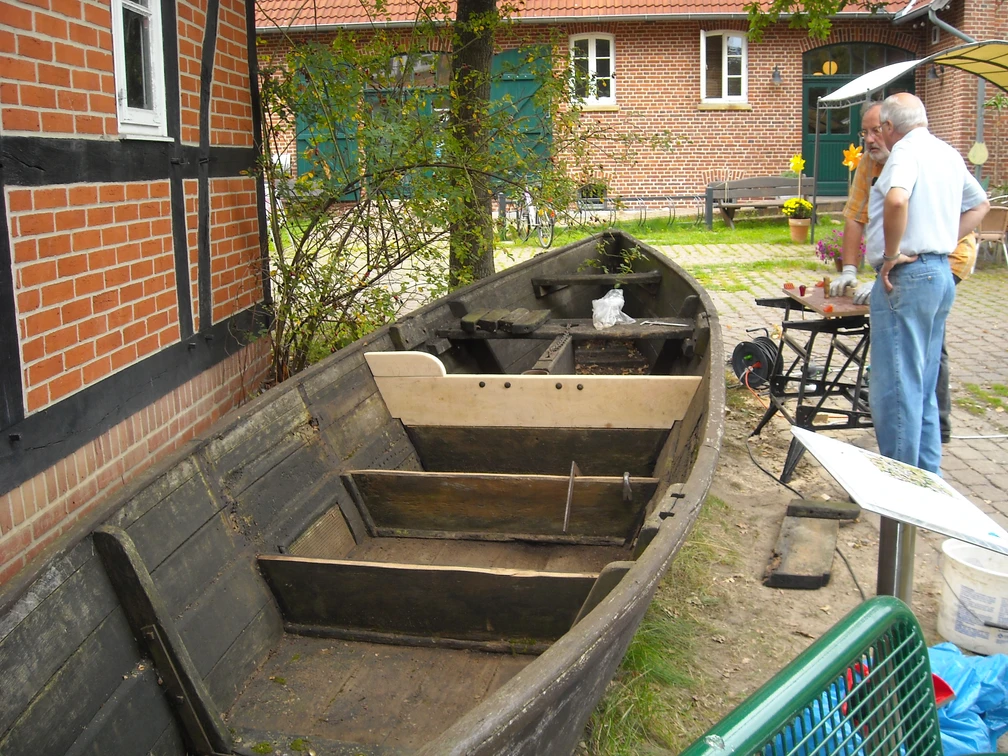 Das Bild zeigt ein altes, teilweise restauriertes Holzboot auf einem Platz vor einem Backsteingebäude.