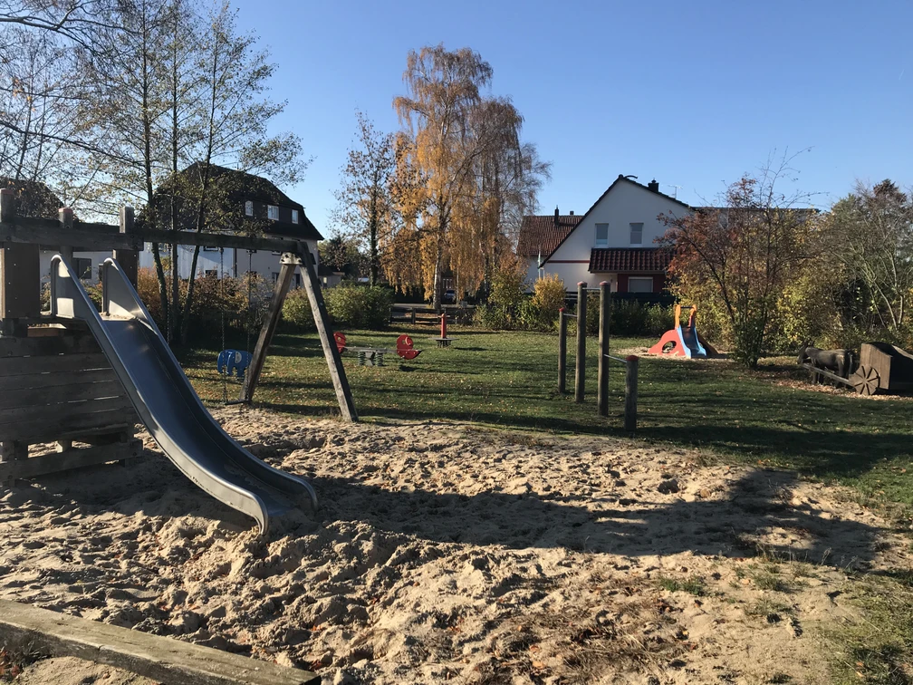Ein moderner Spielplatz mit Klettergerüst, Rutsche und Wippe, eingerahmt von herbstlichen Bäumen.