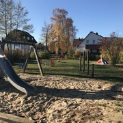 Ein moderner Spielplatz mit Klettergerüst, Rutsche und Wippe, eingerahmt von herbstlichen Bäumen.