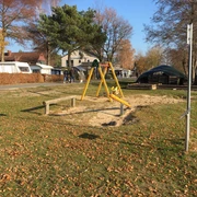 Spielplatz Lüttjen Mardorf Spielplatz mit Schaukel und Sandkasten auf einer sonnigen Wiese, umrahmt von Bäumen und Wohnhäusern.