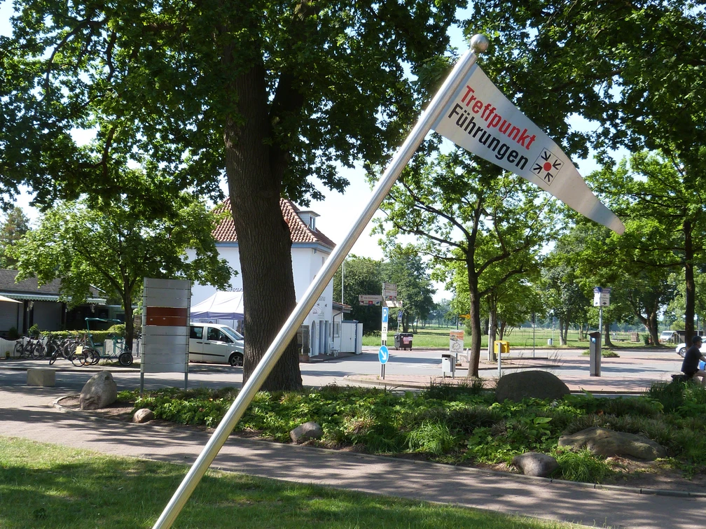 Treffpunkf für Gästeführungen Eine Fahne mit der Aufschrift "Treffpunkt Führungen" neigt sich neben einem Weg in einem grünen Parkbereich.