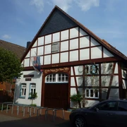 Steinhuder Museen Das Bild zeigt ein traditionelles Fachwerkhaus mit einem steilen Giebel und hölzernen Verzierungen, das Teil des Steinhuder Museums ist. Vor dem Gebäude sind Fahrradständer und ein Baum sichtbar, während rechts ein geparktes Auto zu sehen ist. Der Himmel ist leicht bewölkt.
