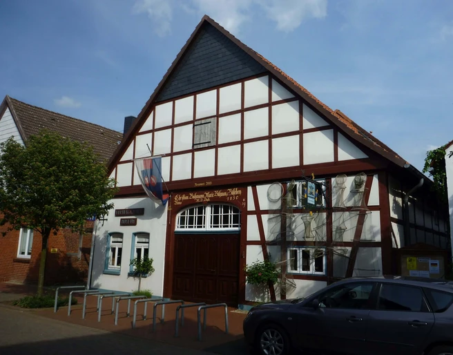 Steinhuder Museen Das Bild zeigt ein traditionelles Fachwerkhaus mit einem steilen Giebel und hölzernen Verzierungen, das Teil des Steinhuder Museums ist. Vor dem Gebäude sind Fahrradständer und ein Baum sichtbar, während rechts ein geparktes Auto zu sehen ist. Der Himmel ist leicht bewölkt.