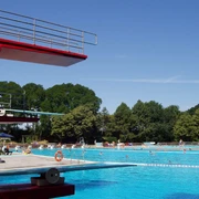 Freibad Bokeloh Großes Freibad mit Sprungturm und Becken, umgeben von Bäumen und sommerlich blauem Himmel.