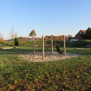 Spielplatz mit Klettergerüst auf Sand, grüner Wiese und Rutsche, umgeben von herbstlichen Bäumen.