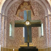 mandelsloh-st.osdag-kirche-kreuz-altar Ein großes Steinkreuz auf einem Sockel im Vordergrund, dahinter verzierte Wand mit gemalter Engelsfigur.