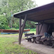 Grillhütte Hagenburg mit Auswanderer Grillhütte aus Holz mit überdachtem Sitzbereich und einem Auswandererschiff im grünen, bewaldeten Hintergrund.