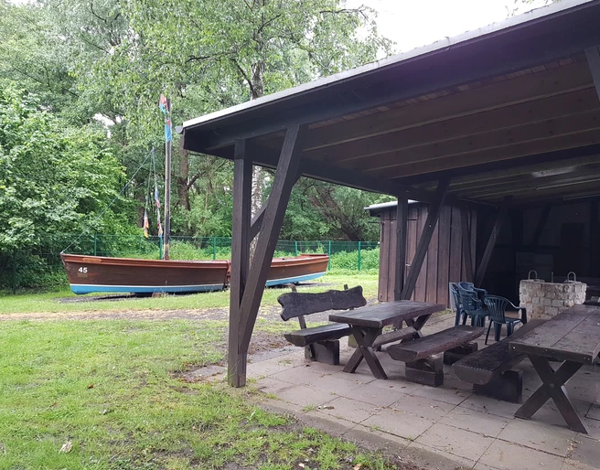 Grillhütte Hagenburg mit Auswanderer Grillhütte aus Holz mit überdachtem Sitzbereich und einem Auswandererschiff im grünen, bewaldeten Hintergrund.