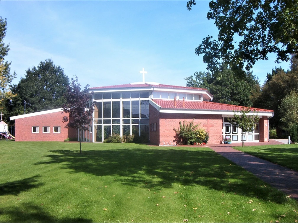 Freikirche_Augustfehn Moderne Freikirche mit rotem Ziegeldach und großem Glasfenster, eingebettet in eine grüne Landschaft.