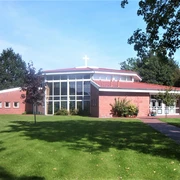 Freikirche_Augustfehn Moderne Freikirche mit rotem Ziegeldach und großem Glasfenster, eingebettet in eine grüne Landschaft.