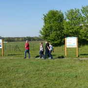 Trinkwasser-Lehrpfad Menschen spazieren auf einem Lehrpfad entlang Infotafeln, umgeben von einer weitläufigen, grünen Landschaft.