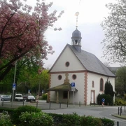Eine kleine Kapelle mit Schindeldach und Turm, umgeben von blühenden Bäumen und Straßenpflaster.