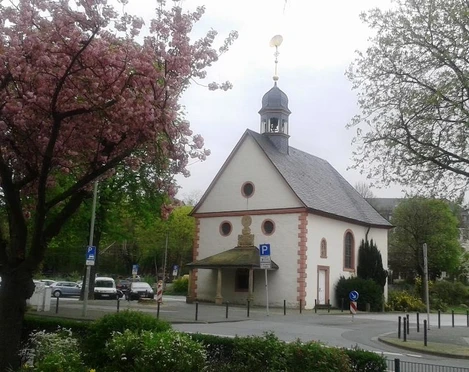 Eine kleine Kapelle mit Schindeldach und Turm, umgeben von blühenden Bäumen und Straßenpflaster.