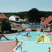 Kinderbecken mit Rutsche und Wasserspielen vor einem Waldschwimmbad an einem sonnigen Tag.