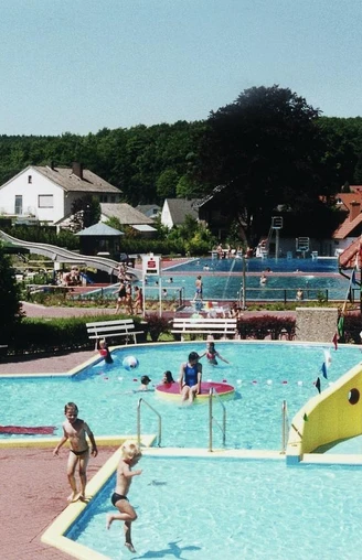 Kinderbecken mit Rutsche und Wasserspielen vor einem Waldschwimmbad an einem sonnigen Tag.