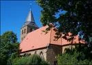 Backsteinkirche mit rotem Dach und spitzem Turm, umgeben von Bäumen unter strahlend blauem Himmel.