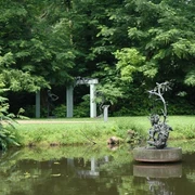 Bronzeskulptur auf Teich mit grünen Bäumen und einer Gartenlaube im Hintergrund.