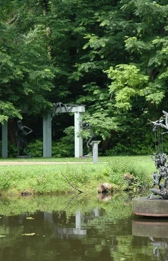 Bronzeskulptur auf Teich mit grünen Bäumen und einer Gartenlaube im Hintergrund.