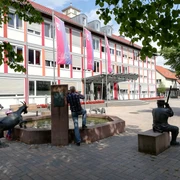 Mensch und Kind betrachten Ziegen am Brunnen vor modernem Gebäude, harmonisch in Lügdes Zentrum.